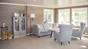 Un salon moderne avec un canapé gris, deux fauteuils, une table basse et un meuble avec des portes en verre. De grandes fenêtres laissent entrer la lumière naturelle et offrent une vue sur la verdure environnante.