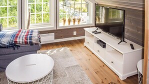 Un salon moderne avec une grande télévision, un plancher en bois, un canapé gris et une fenêtre agrémentée de plantes en pot.