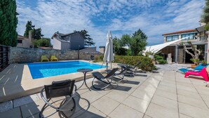Ein Schwimmbecken mit Liegestühlen und einem Rollstuhl, umgeben von Steinmauern und Grünpflanzen.
