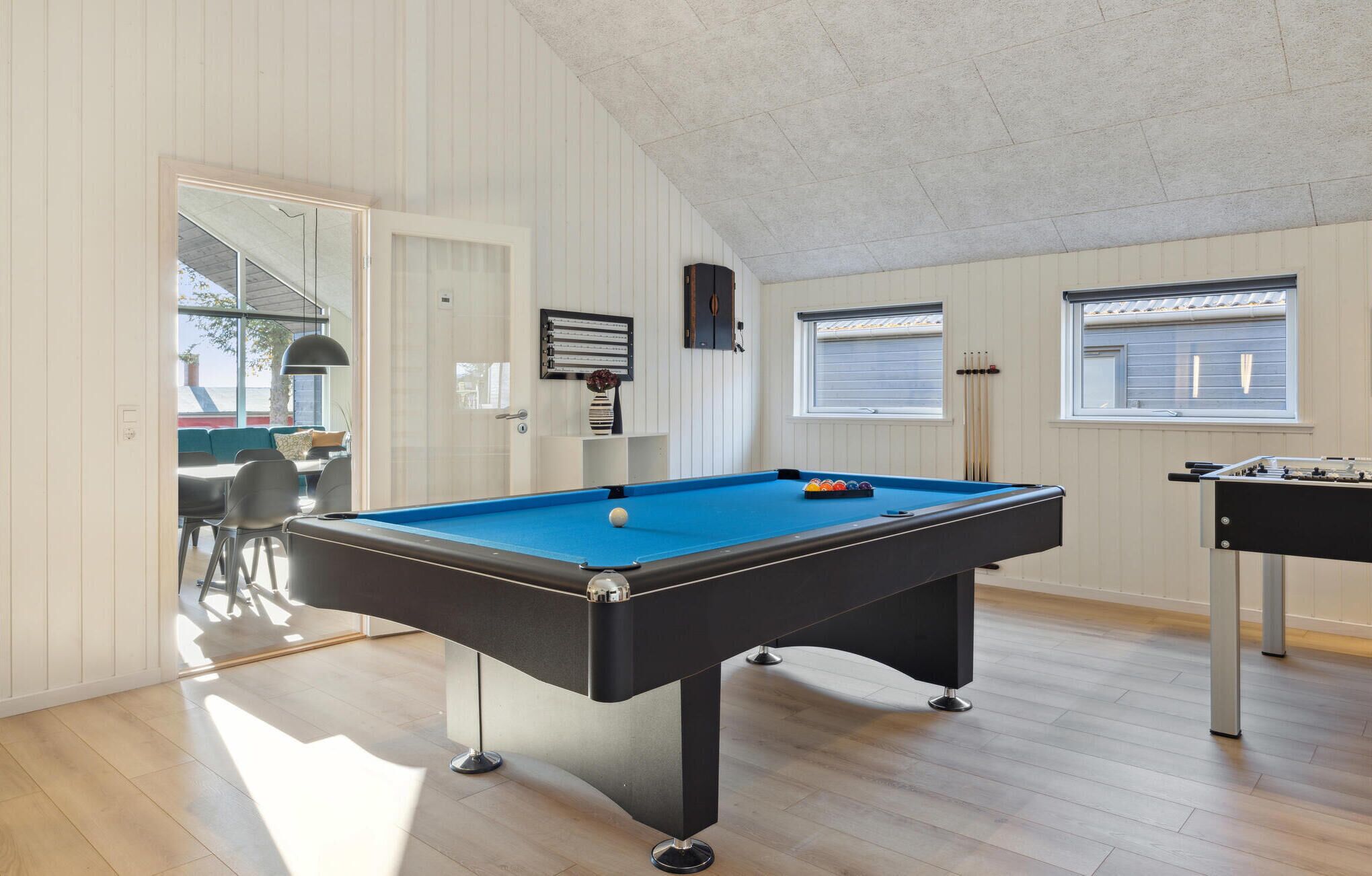 A pool table in a room with wooden flooring and white walls, a door leading outside, and a window with blinds.