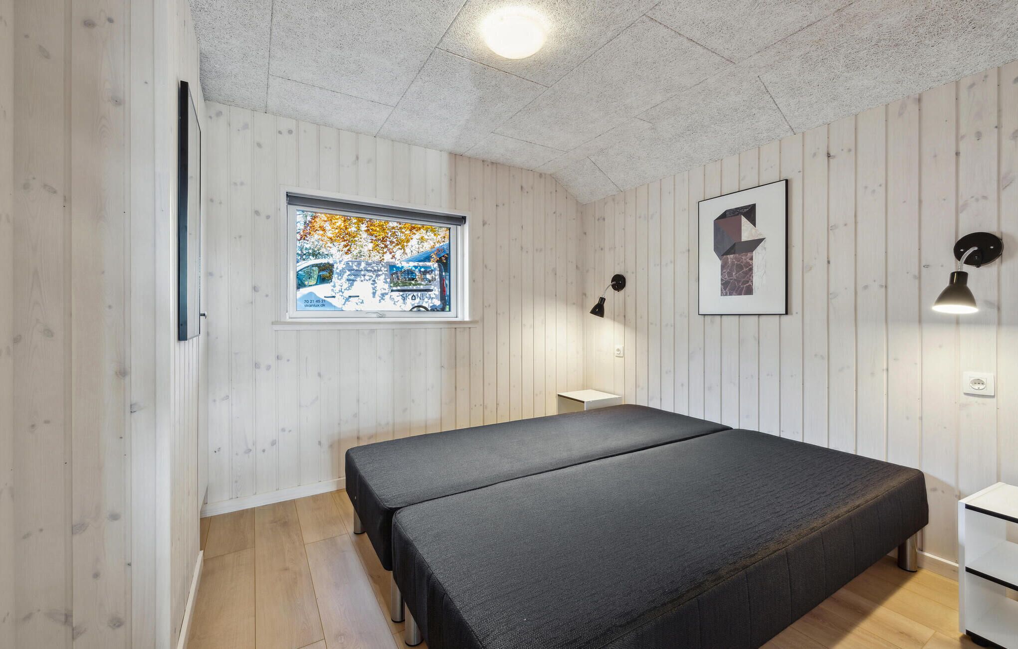 A room with a bed, a window with a view of a street, a wall-mounted lamp, and a framed picture.