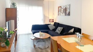Ein modernes Wohnzimmer mit einem dunkelblauen Sofa, einem Glastisch und einem Holzespeisetisch mit Stühlen. Es gibt ein großes Fenster mit durchsichtigen Vorhängen, einen Flachbildfernseher und ein Gemälde an der Wand.