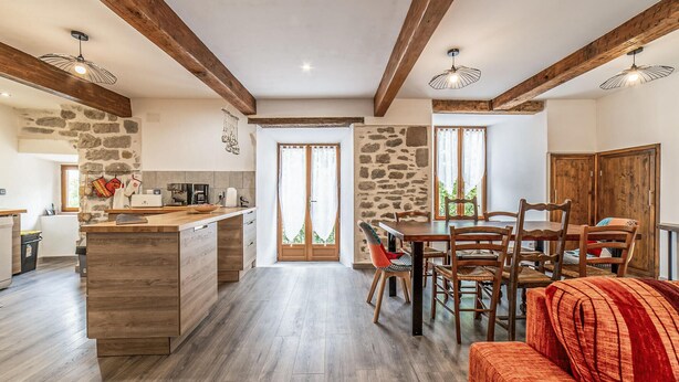 Una cocina amplia con una isla central, vigas de madera, paredes de piedra y un comedor con sillas y mesa de madera.