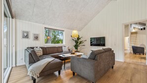 A modern living room with a high ceiling, a large window with a view of trees, a flat-screen TV mounted on the wall, and a cozy seating area with a sofa and armchairs. - Cozy home in Krakow am See with sauna (Krakow am See)
