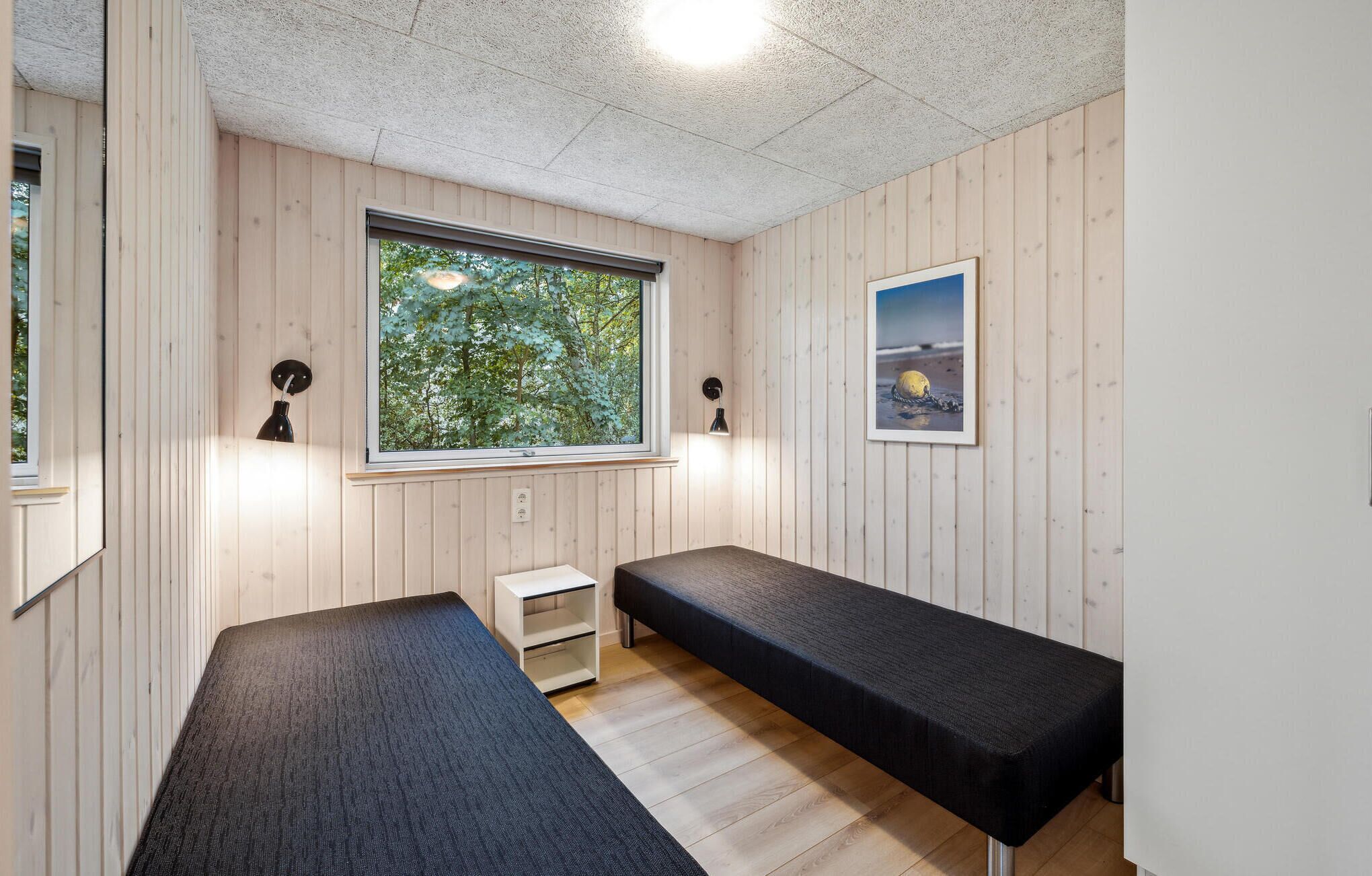 Une pièce aux murs en bois, avec une grande fenêtre donnant sur des arbres, un banc et deux lampes fixées au mur.