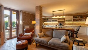 A modern living room with a sofa, armchair, and ottoman, a dining area with a table and chairs, and a kitchen with brick walls and modern appliances. - Lovely home in Castiglione Del Lago (Castiglione Del Lago)