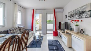 Una sala moderna con sofá, mesa de comedor y televisor. Tiene un balcón con cortinas rojas y un cuadro de un paisaje urbano en la pared.