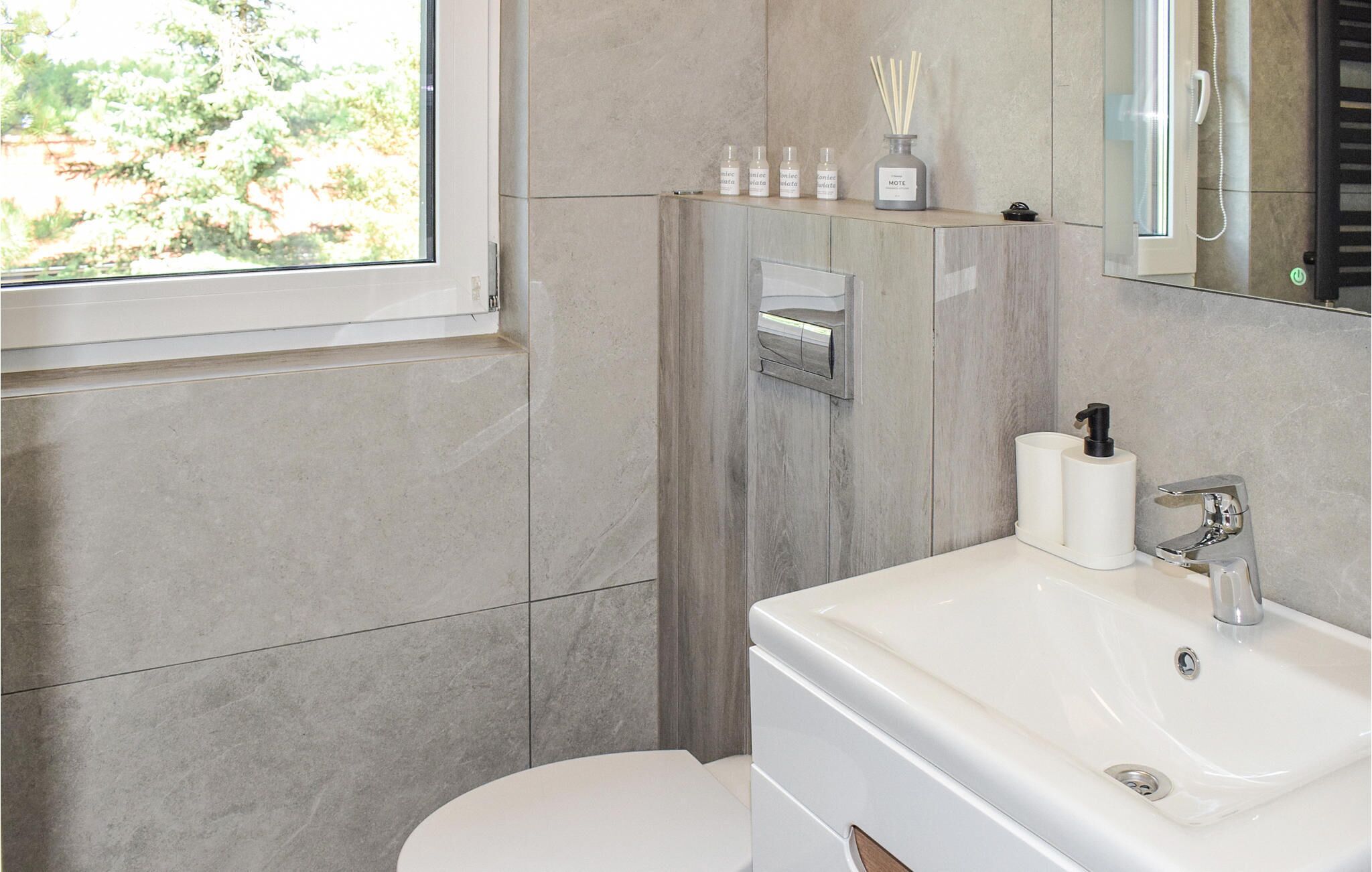A modern bathroom with a toilet, sink, and shower. There are toiletries on a shelf and a mirror above the sink.