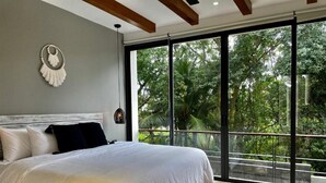 A bedroom with a large bed, a view of lush greenery through floor-to-ceiling windows, and a wooden ceiling beam. - BT Rooms Tulum (Tulum)