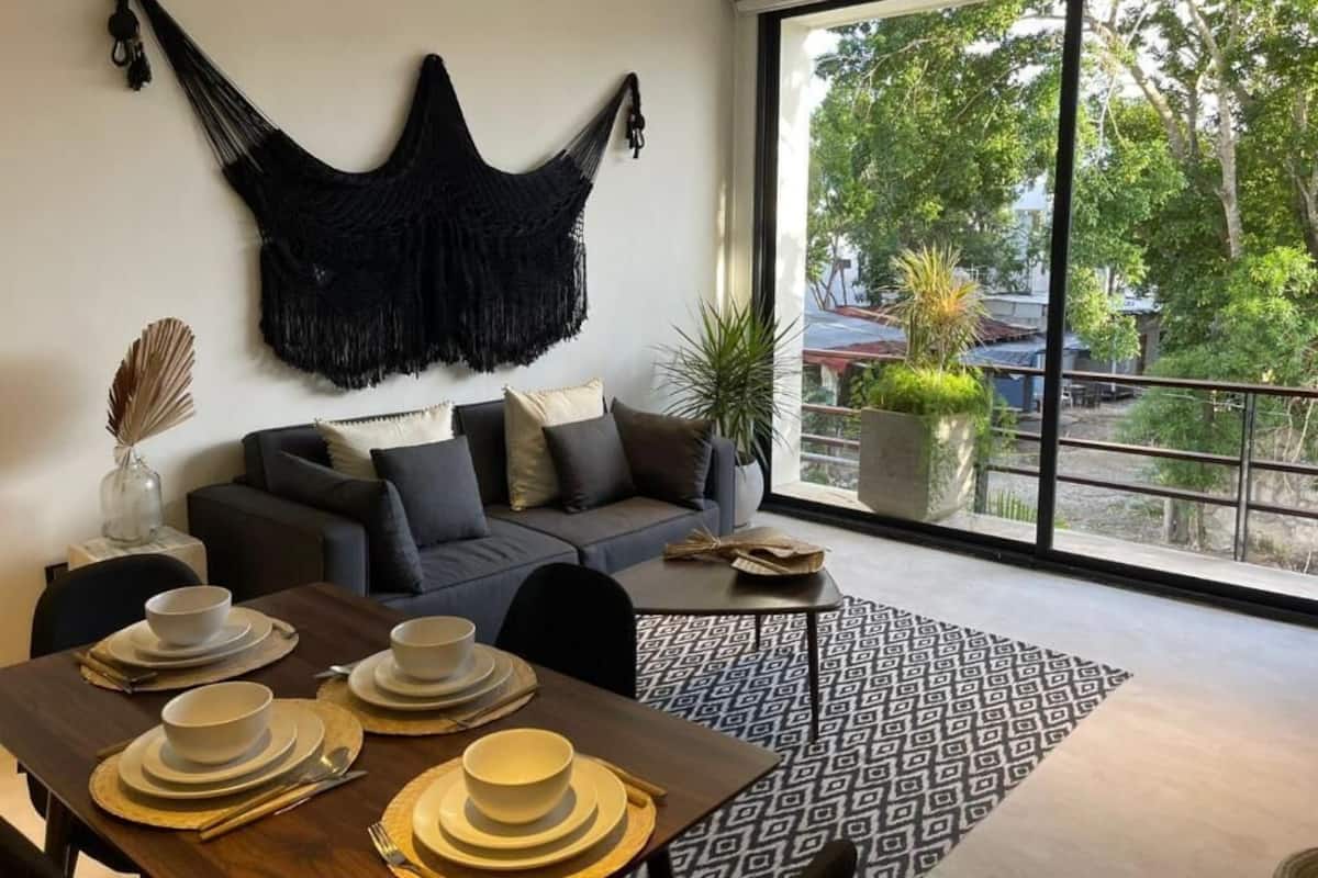 Ein modernes Wohnzimmer mit großem Fenster, einem schwarzen Wandbehang aus Makramee, einem grauen Sofa und einem Essbereich mit Geschirr und Besteck.