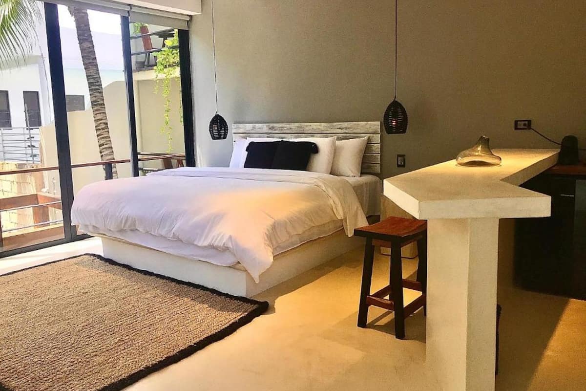 Ein Schlafzimmer mit einem großen Bett, einem Schreibtisch und einem Stuhl. Ein Fenster bietet Blick auf Palmen und ein Gebäude.