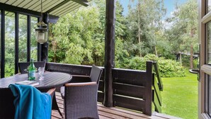 Uma área de estar coberta ao ar livre com mesa e cadeiras, uma garrafa de S.PELLE e vista para um jardim bem cuidado e árvores.
