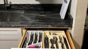 Una cocina con cubierta negra, armarios blancos y un cajón con utensilios.