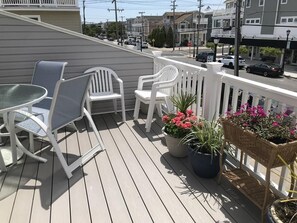 A deck with white chairs and a table, potted plants, and a view of the street.