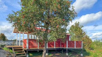 A red wooden deck with a staircase leading up to a covered area, surrounded by lush greenery and a clear sky.
