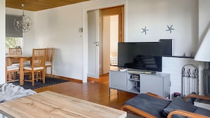 Uma sala de estar moderna com uma mesa de jantar de madeira, um sofá, uma TV em um armário cinza e uma porta que dá para outro quarto.