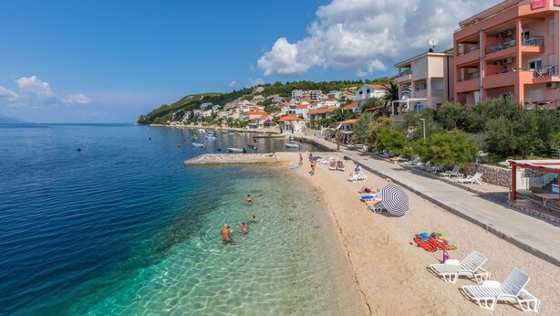 A beach with clear water, sun loungers, umbrellas, and a row of buildings along the shore. - Luxurious home in Komarna (Komarna)