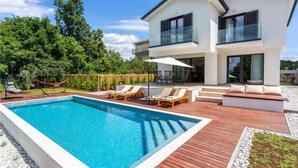 Ett modernt hus i två våningar med en swimmingpool, utomhusmöbler och utsikt över den omgivande naturen.