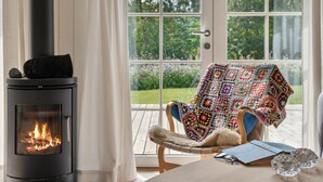 A cozy room with a wood-burning stove, a chair with a colorful blanket, and a view of a garden through large windows. - Amazing home in Gilleleje with WiFi (Gilleleje)