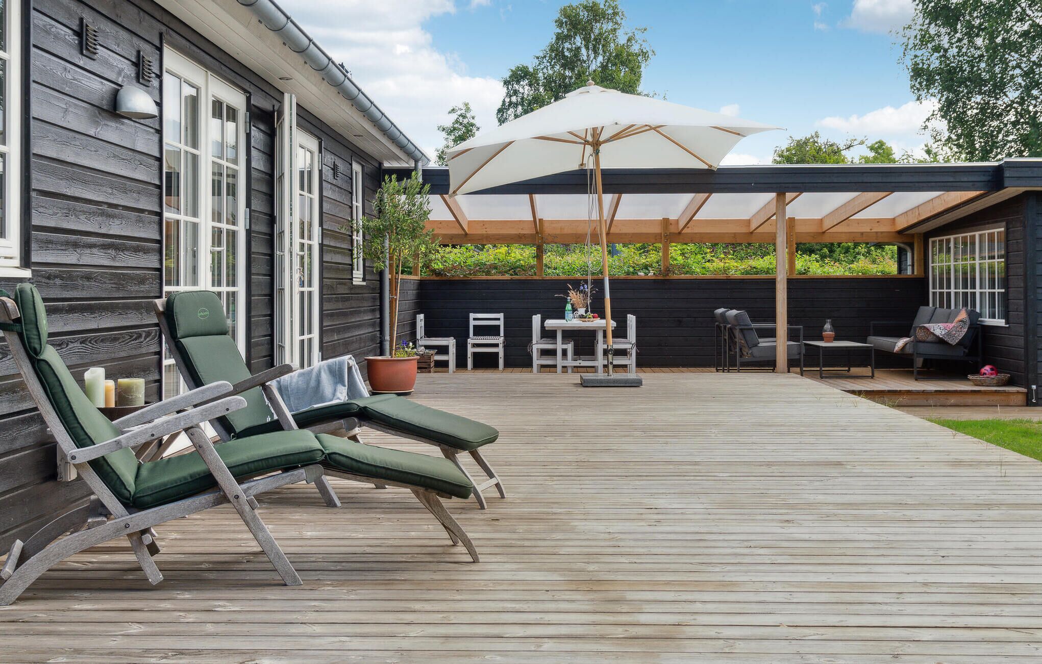A wooden deck with lounge chairs and a table, a covered patio area with chairs and a table, and a building with large windows.
