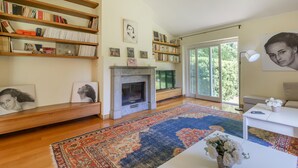 Een ruime woonkamer met een open haard, boekenkasten, een groot vloerkleed en uitzicht op groen via de schuifdeuren.