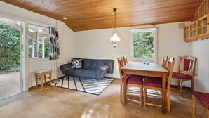 A cozy room with a wooden ceiling, a grey sofa, a dining table with chairs, and large windows offering a view of greenery.