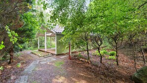 A small wooden structure with a roof, surrounded by dense green foliage and a stone path leading to it.