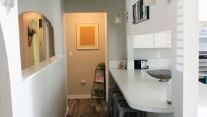 Un couloir étroit, doté d’un plancher en bois, mène à une cuisine avec un comptoir blanc, des tabourets de bar et des luminaires fixés au mur.