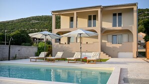 Ein zweistöckiges Gebäude mit Pool, Außenbereich mit Sonnenschirmen und einer Hanglage im Hintergrund.