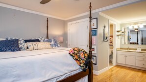 A bedroom with a four-poster bed, a wooden floor, a bathroom with a vanity and mirror, and a door leading to another room.