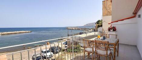A balcony with a table and chairs overlooking a coastal area with parked cars and a rocky breakwater.