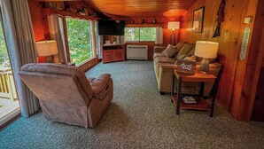 Ein gemütliches Blockhauss-Interieur mit Wohnbereich, einer Couch, einem Sessel, einem Tisch mit Lampe und einem Fernseher.