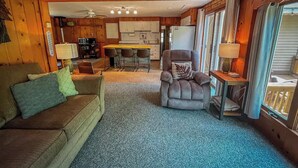Ein gemütliches Blockhaus-Interieur mit Holzbalkendecke, einem Essbereich, einem Wohnzimmer mit Sofa und Sesseln sowie einer Küche im Hintergrund.