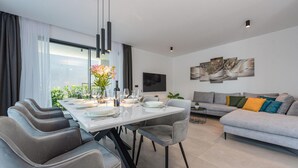 A modern living room with a grey sofa, a dining table set for four, and a large window with curtains. - Beach front home in Pasman (Pasman)