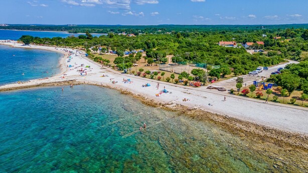 Vista aérea de una playa con aguas cristalinas, una orilla arenosa y una mezcla de vegetación y edificios al fondo.