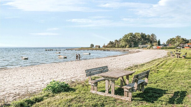 Une plage avec un banc en bois et une table de pique-nique, des bateaux sur l’eau et des maisons en arrière-plan.