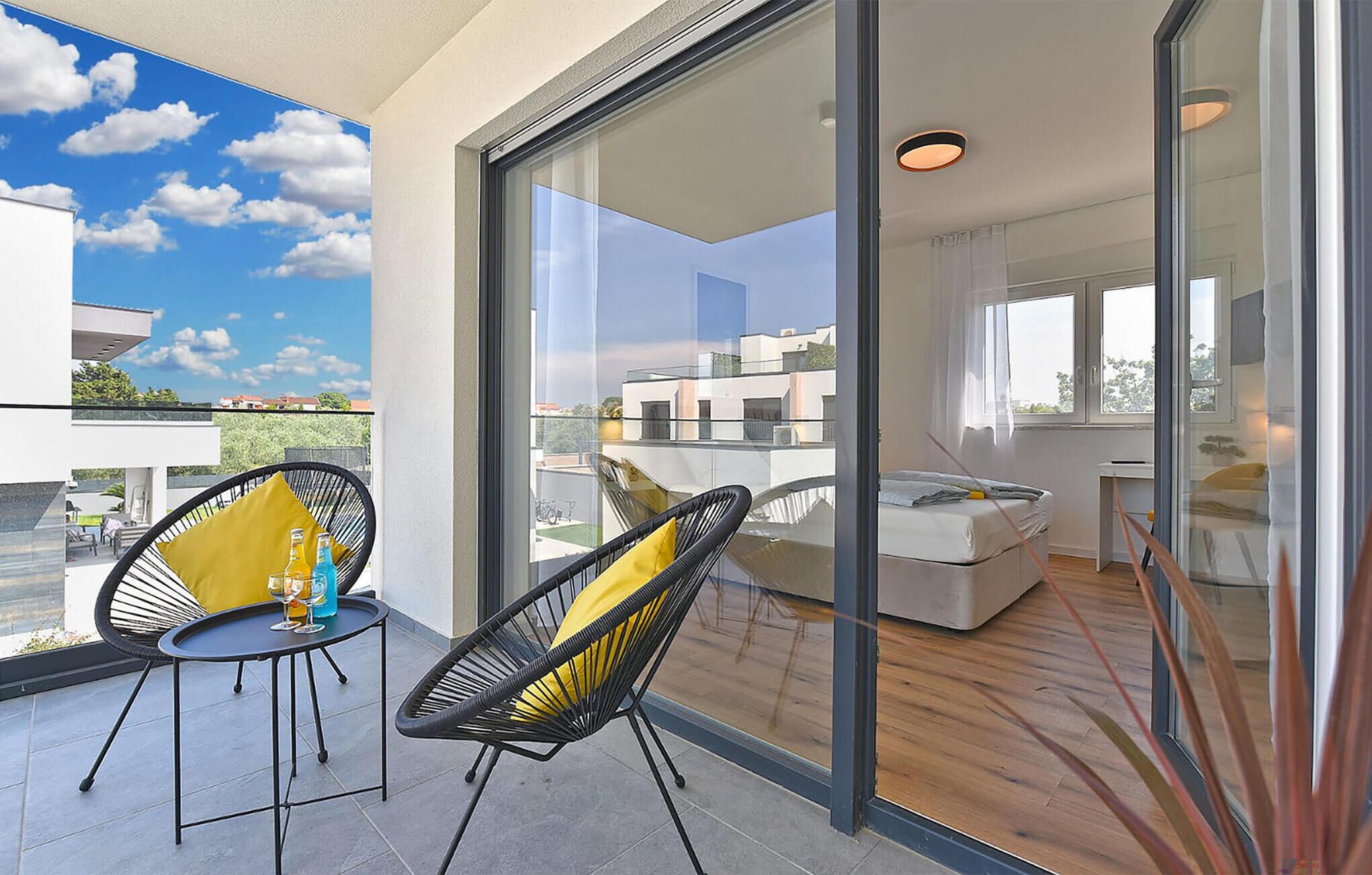 A balcony with two chairs, a table, and a glass door leading to a bedroom with a bed and a desk.