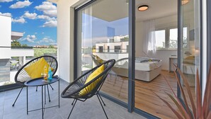 Varanda com duas cadeiras, uma mesa e porta de vidro que dá para um quarto com cama e escrivaninha.