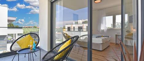A balcony with two chairs, a table, and a glass door leading to a bedroom with a bed and a desk.