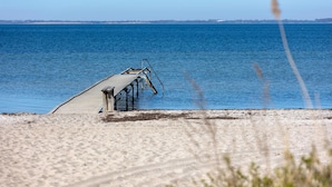 Un muelle de madera que se adentra en una masa de agua tranquila, con una playa de arena en primer plano y una orilla lejana.