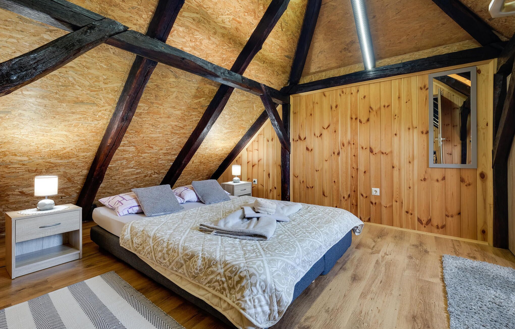 Um quarto com teto inclinado, paredes de madeira, uma cama com roupa de cama estampada, mesas de cabeceira com luminárias e um espelho na parede.