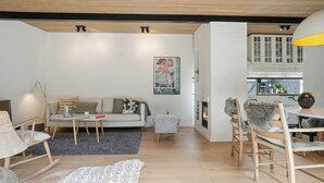 Una sala de estar moderna con sofá, mesa de centro y sillones, chimenea y área de cocina con armarios y mesa de comedor.