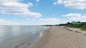 Una amplia playa de arena con agua tranquila, algunas personas caminando y casas al fondo.