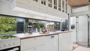 Cocina moderna con armarios blancos, horno integrado, cafetera y vista al jardín.