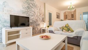Een woonkamer met een flatscreen televisie op een witte tv-meubel, een witte salontafel met een schaal snacks, een grijze bank met decoratief kussen, een groot wanddecor en een groene kroonluchter.