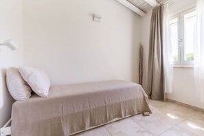 A single bed with a beige bedspread in a room with white walls, a window with curtains, and a white floor lamp.