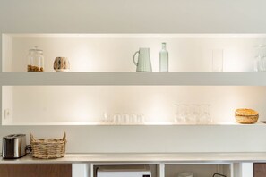 A kitchen with two white shelves, one holding a jar, a mug, a pitcher, and a bottle, and the other with glasses and a basket.