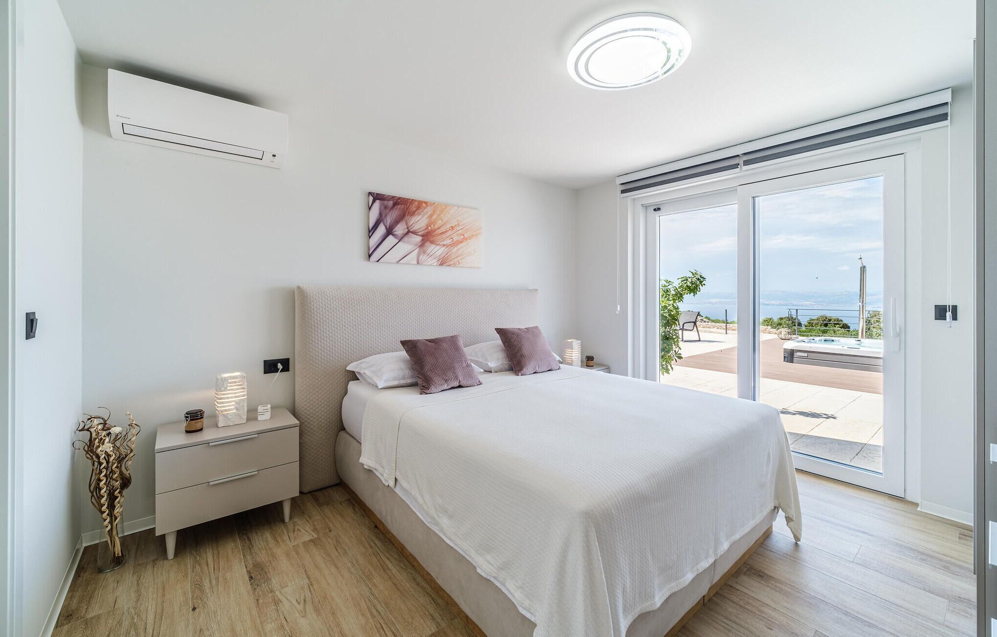A bedroom with a bed, nightstands, a wall-mounted air conditioner, a painting, and a view of the outdoors.