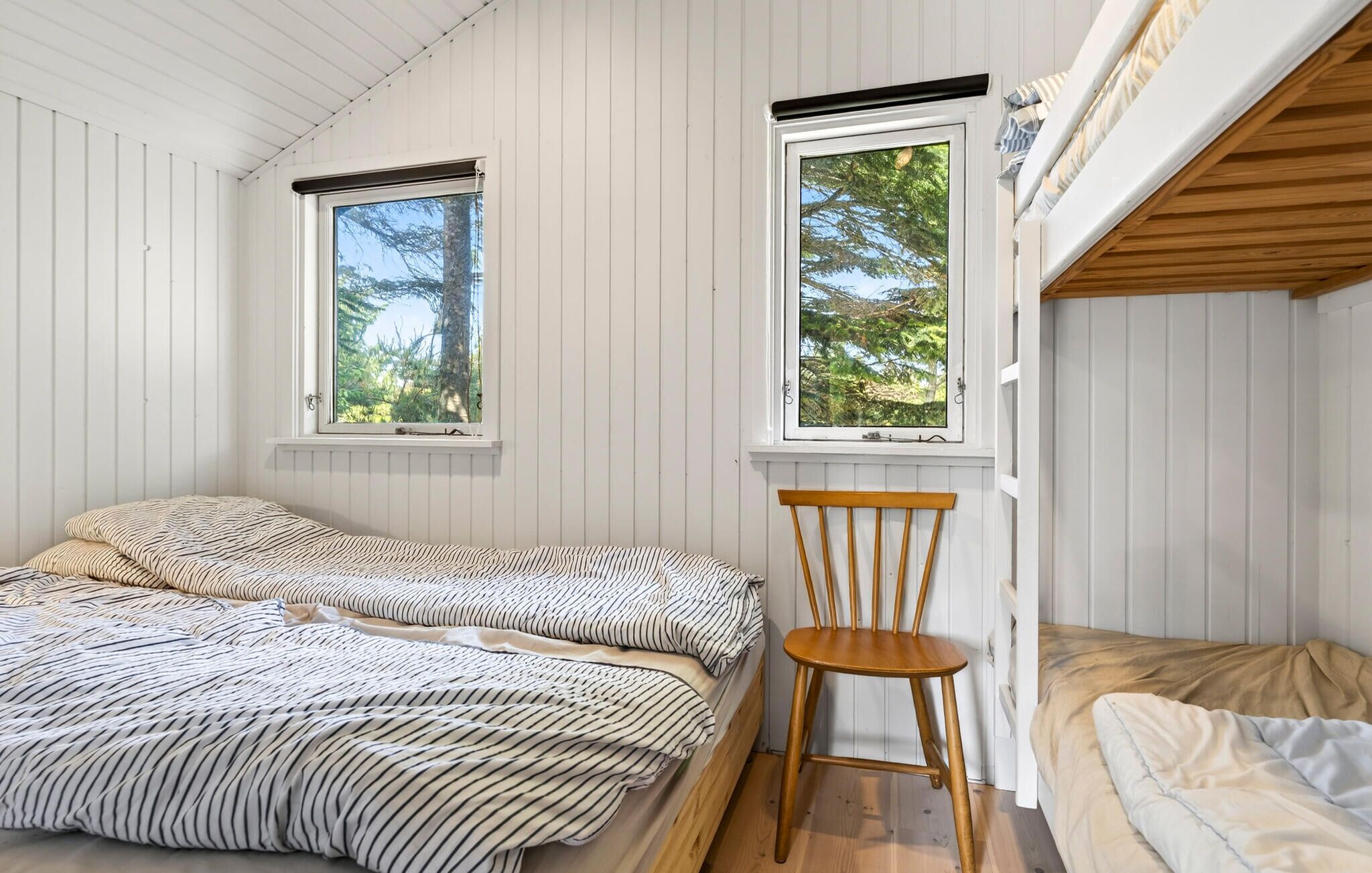 Une chambre avec deux lits, une chaise en bois et deux fenêtres donnant sur des arbres.