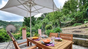 Une table en bois, dressée pour deux personnes, est placée sous un parasol blanc. Elle est entourée de verdure et offre une vue sur un paysage vallonné.